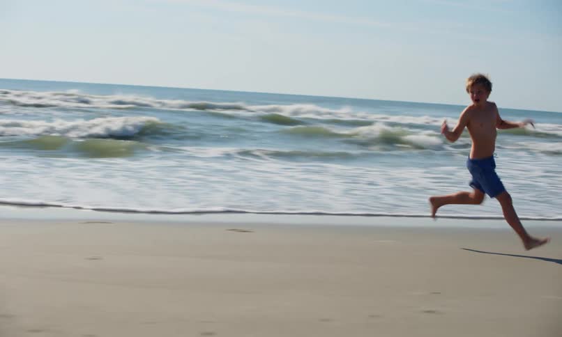 A boy runs ahead as two children happily play chase on the shoreline as the surf rolls in on a sunny day at the beach.