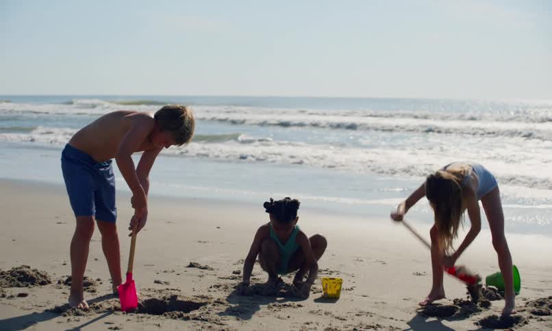 Three kids, a boy and two girls, use play shovels and plastic buckets to dig holes and build sand castles on a sunny day at the beach.