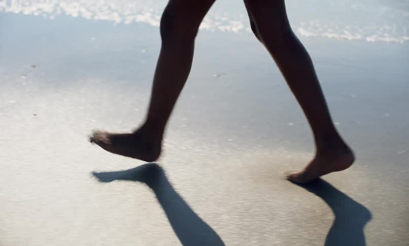 A young black girl child creates a shadow as she walks with toes in the sand along the shoreline of a beach as the surf flows in.