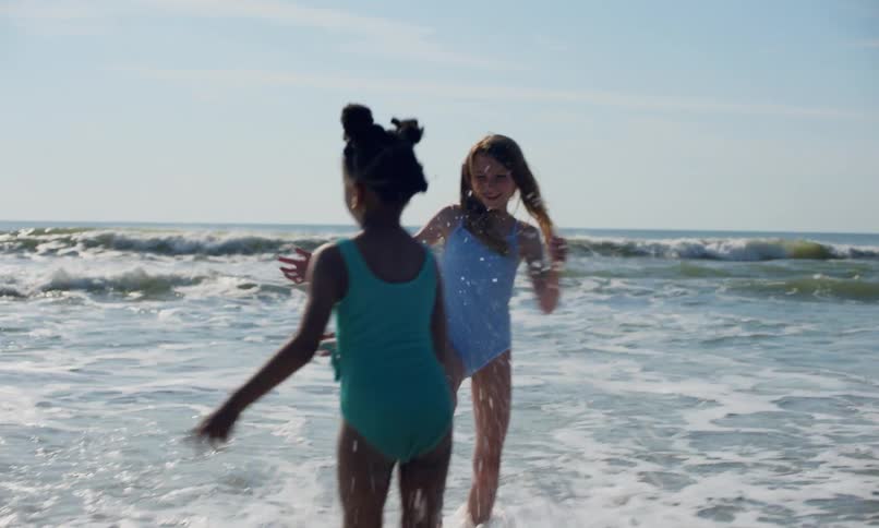 Three diverse children play in the waves on the shoreline by the ocean under a blue sky on a sunny day.