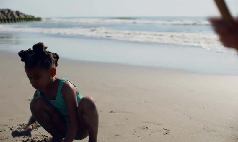 A young girl of color plays in the sand at the beach next to a young blonde girl who is digging with a shovel as the sun glints off the waves washing in the surf behind them.