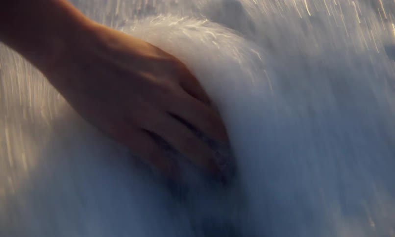 Close up of water on the beach shoreline with a female's hand reaching down to grab a piece of translucent blue seaglass as a wave brings the surf in