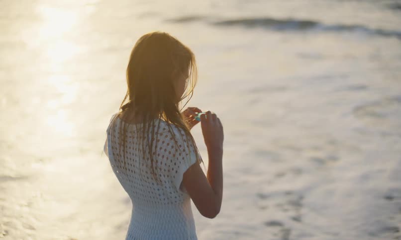 A girl in a white coverup dress stands with damp hair on the ocean shore letting the waves touch her feet at sunset while examining a piece of blue glass in her hands