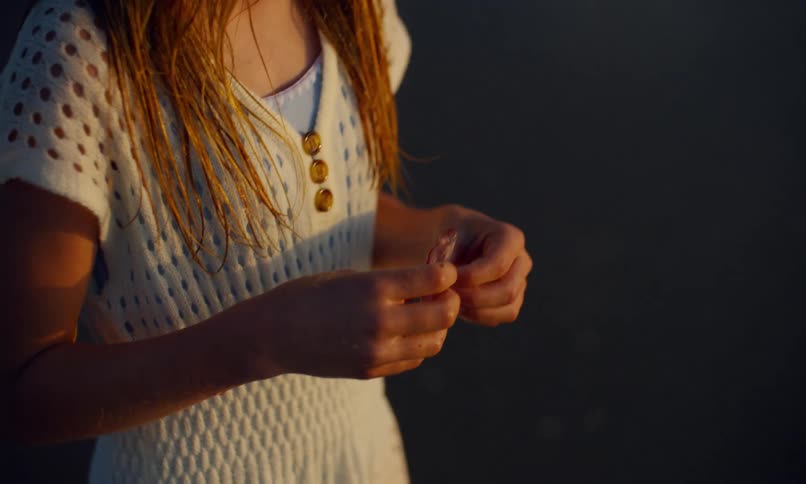 A young girl with long hair and wearing a white dress picks up and admires a piece of translucent sea glass from the surf on a beach at sunset.