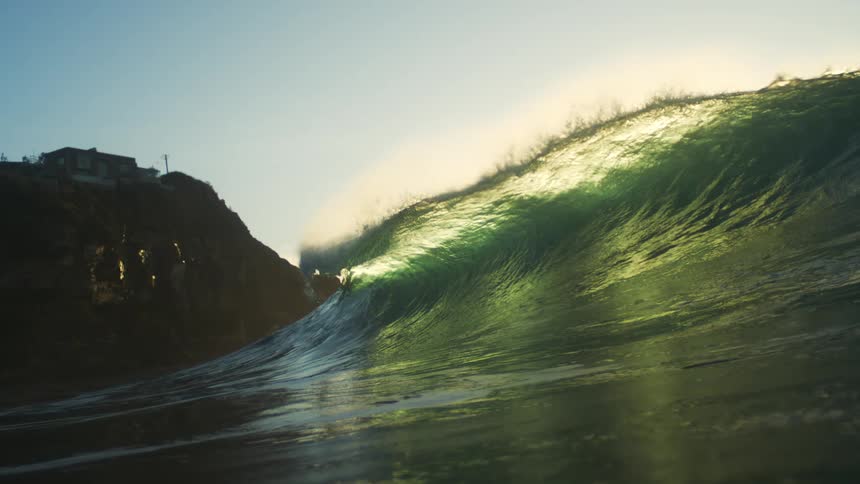 A large ocean wave is illuminated by sunlight with a rocky cliff and a house visible in the background