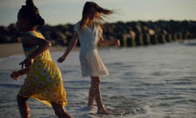 Slow motion of a happy young black girl child splashing water behind her and running past another young girl as they play along the seashore at sunset.