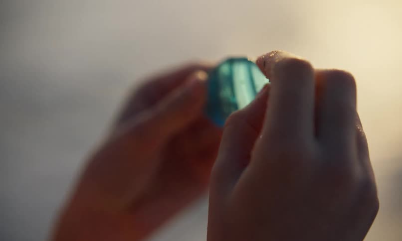 A young girl with long hair holds a translucent blue piece of textured decorative glass in both hands and thoughtfully examines it with her fingers as she holds it up to the light of the setting sun.