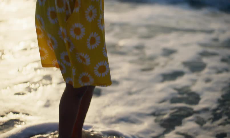 A young black girl child in a yellow, flowered dress happily plays on the sea shore at sunset and opens her mouth with gleeful joy as the waves flow against her bare feet.