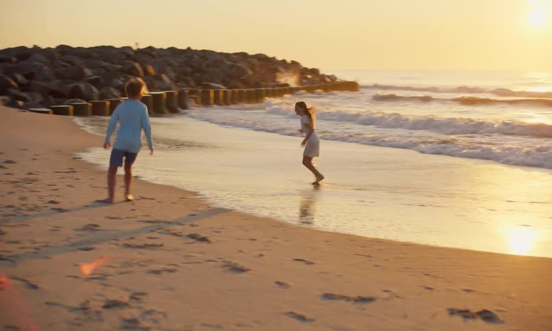 Three children are playing chase with each other on the shoreline of a beach at sunset as waves crash against the sand and the suns rays blur the horizon.