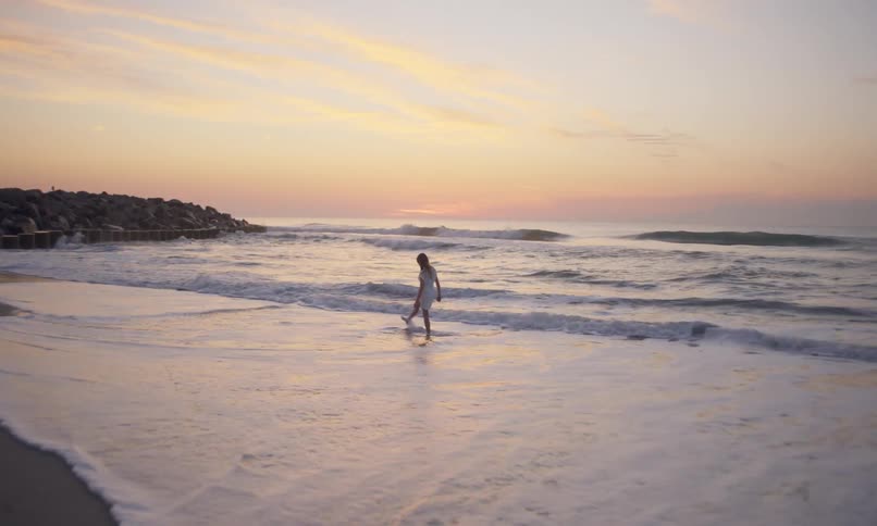 A lone little girl in a white dress walks timidly along the wet, sandy shoreline of a beach looking out at the muted light on the horizon just after sunset as waves roll in crashing against her legs.
