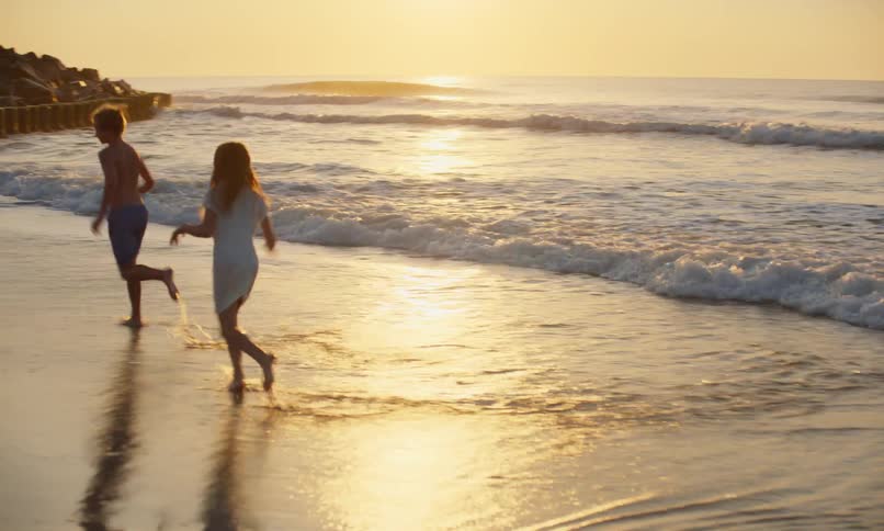 Three children are joyfully playing chase with each other in the shallow waves of the seaside at sunset's golden hour.