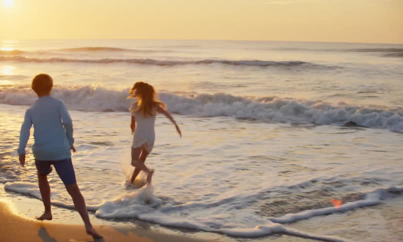 A group of children run and play in the sand and surf of a beach where the golden glow of the setting sun is reflecting off of the incoming waves.