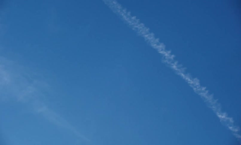 A thin aircraft contrail cuts diagonally across a deep blue sky trailing faint wisps behind it in the clear upper atmosphere