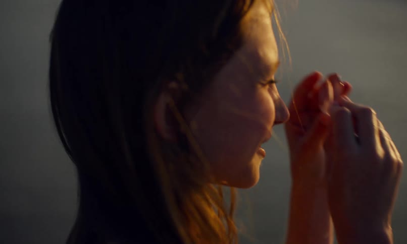 A smiling young girl holds a piece of pink translucent sea glass to her eyes and tries to peer through it while standing on a beach at sunset.