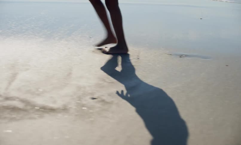 The feet of a young african-american girl walking across wet sand on the shoreline of a beach and casting a sharp shadow in the sunlight