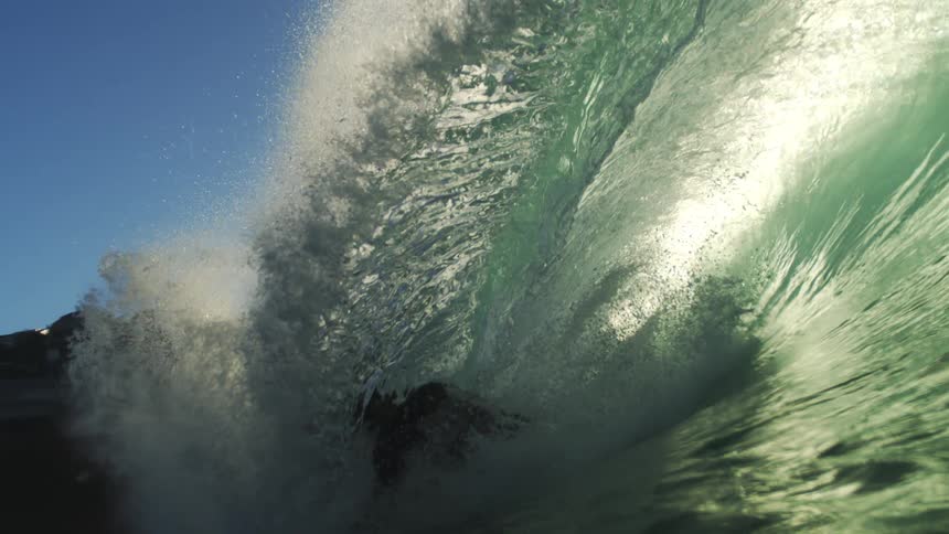 A surfer rides deep inside the barrel of a large sun drenched wave which eventually crashes and sends the view plunging into the murky green depths of the turbulent ocean