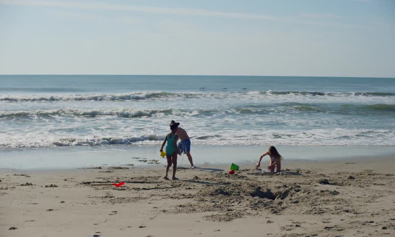 Waves gently roll ashore on a quiet beach as three diverse children happily play in the sand on a sunny day.