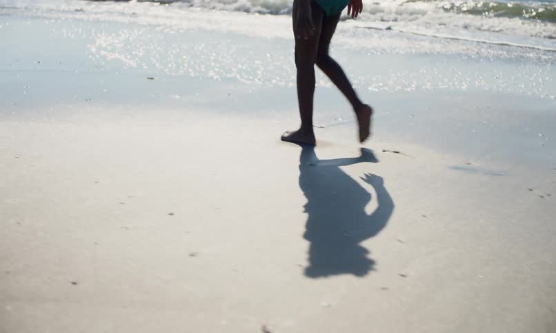 A young, black girl casts a shadow as she walks in her swimsuit barefoot in the wet sand along the shore of the beach on a sunny late afternoon.
