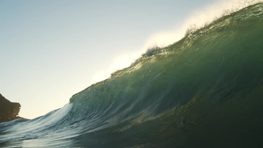 A large backlit ocean wave builds and dramatically crests with the sun flaring behind it before the view plunges underwater into murky green depths