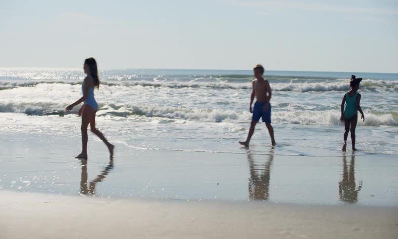Three children playing on the beach with one girl doing a cartwheel as another kicks and runs from the waves coming in and a young boy bends over to pick up something that the surf washed in.