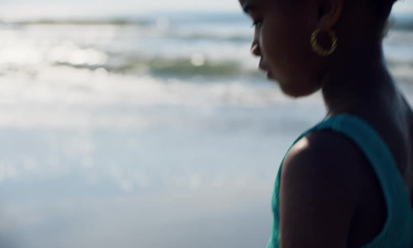 A young black girl in a swimsuit walks quietly along the edge of a beach at midday with waves crashing onto the shore and the sun glinting off the water.