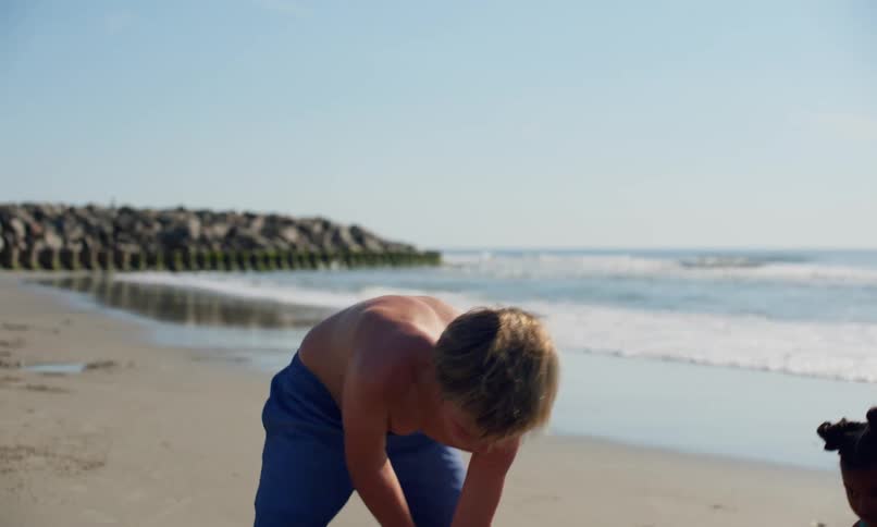 A diverse group of children in swimsuits play with sand at the beach using shovels and sand castle containers as the waves comes ashore behind them.