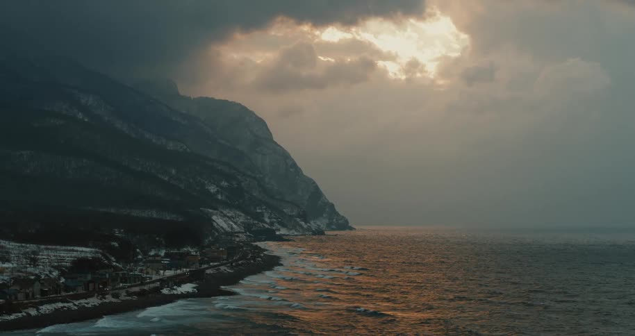 At dawn a coastal town stretches below an elevated viewpoint as cars travel along the winding shore road beneath towering mountains and a soft early light