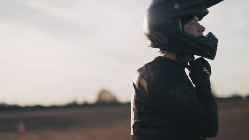 A male motocross rider smiles as he adjusts his safety helmet while standing outdoors