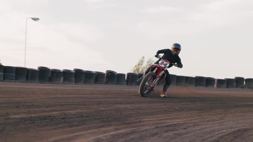 A motorcycle racer wearing a helmet and black suit leans into a turn on a dirt track kicking up dust behind the bike under a bright sky
