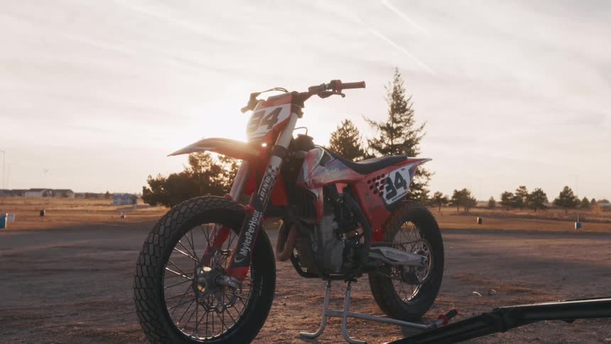 A dirt bike stands parked on its kickstand at the edge of a dusty track glowing in the warm sunset light with long shadows stretching across the ground