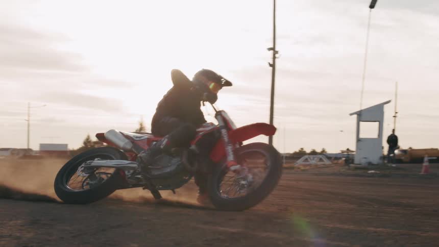 A motorcyclist is performing a sharp turn on a dirt track kicking up dust as the sun sets in the background