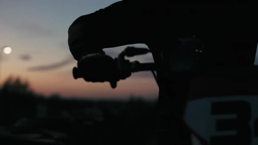 A silhouetted person grips the handlebars of a motorbike seen from the chest up against an orange and purple sunset
