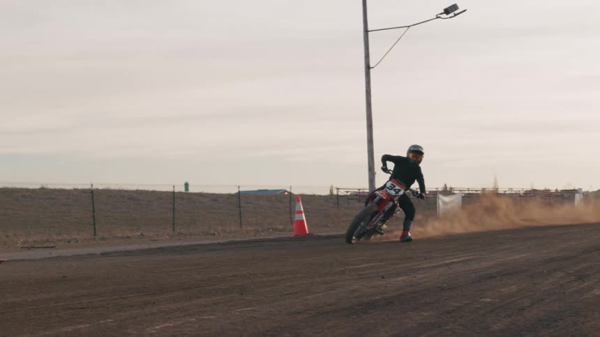 A motorcycle racer wearing a helmet and black suit leans into a turn on a dirt track kicking up dust behind the bike under a bright sky