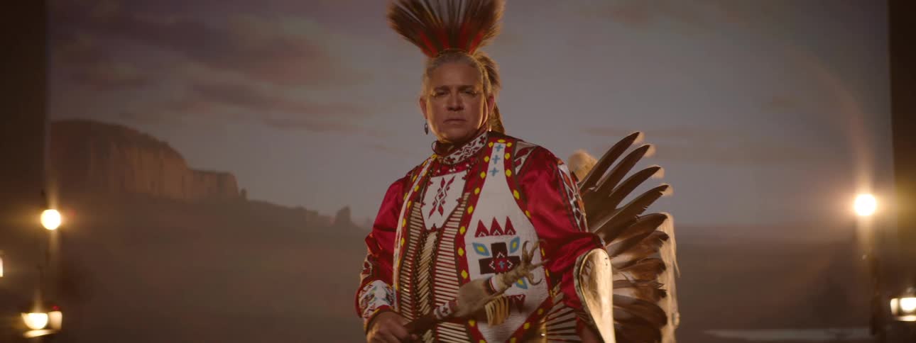 A native american man in traditional regalia stands proudly under warm stage lighting