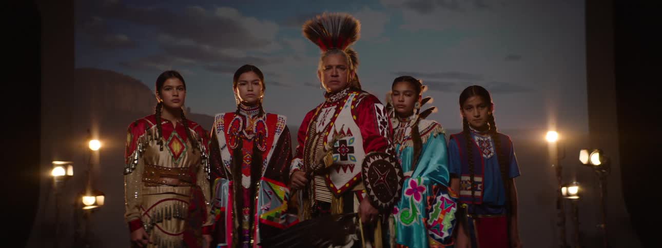 A group of five individuals in traditional native american attire stands together the male prominently featured in an elaborate headdress against a projected outdoor background