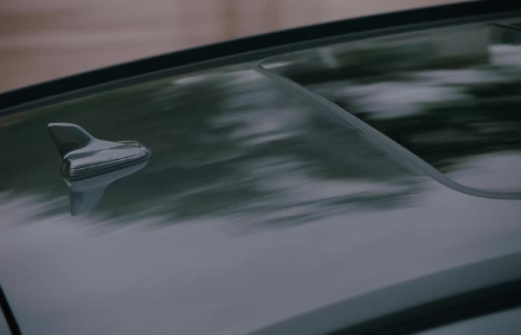 A car is tracked from above while in motion with reflections of passing trees rippling across its glossy sunroof and roof panel
