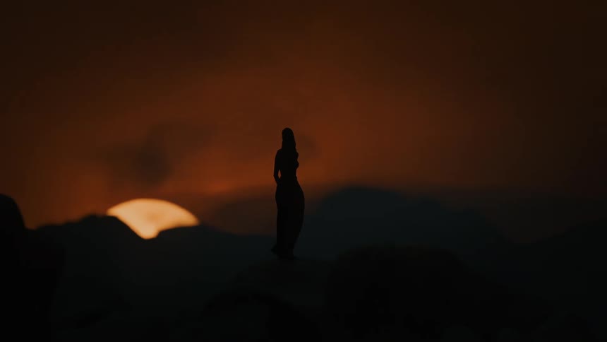 A silhouetted figure stands on a rocky terrain against a dramatic orange sky at sunset