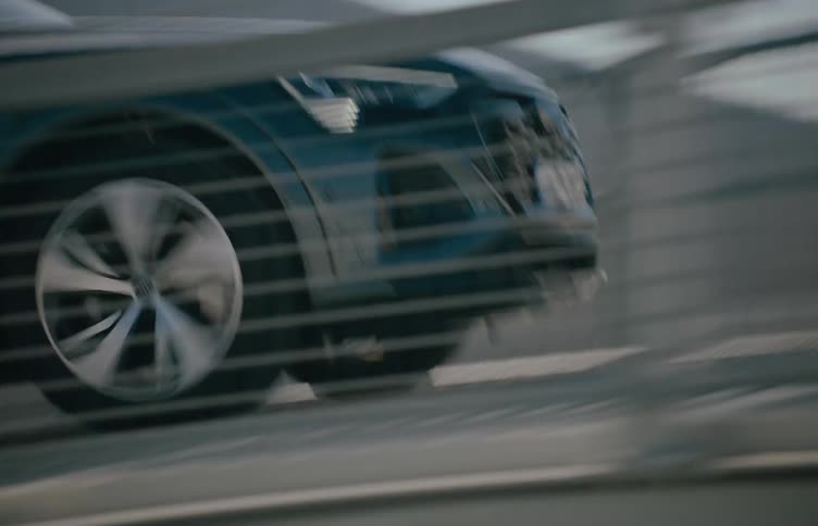 The front of a dark blue car with its headlights visible speeding behind a grey railing up a ramp