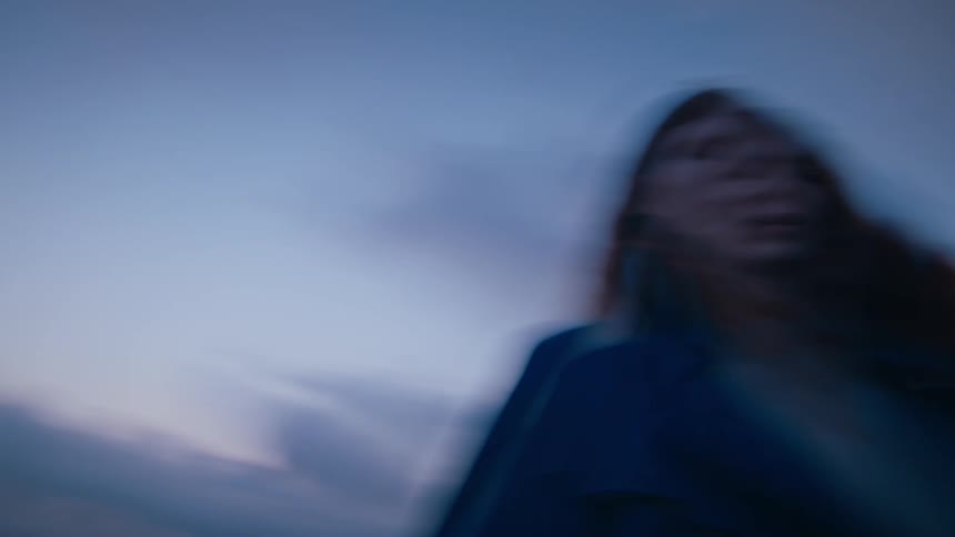 A blurry shot of a woman with long flowing red hair and a dark jacket under a hazy cloudy sky