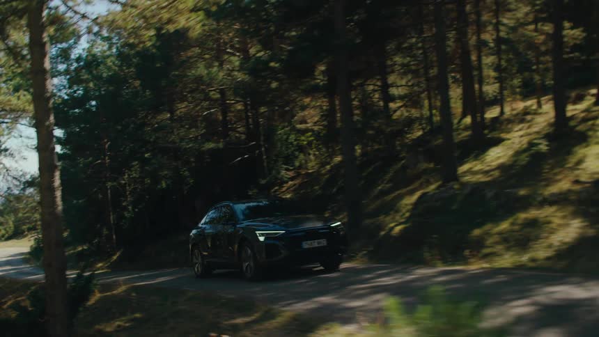 A darkcolored car winds uphill through a forested road as its tracked in motion weaving past sunlit trees and curving through patches of shade and light