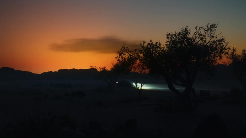 A vehicle with bright headlights drives through a desert road at sunset