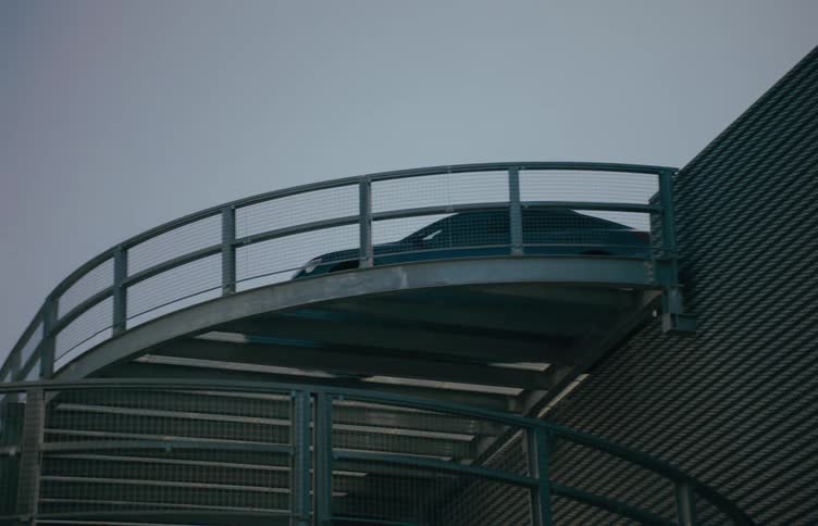 A dark car drives across a section of a curved elevated metal ramp attached to a modern building