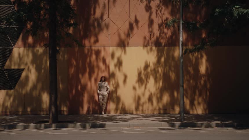 Woman stands in sunlight against wall with tree shadows