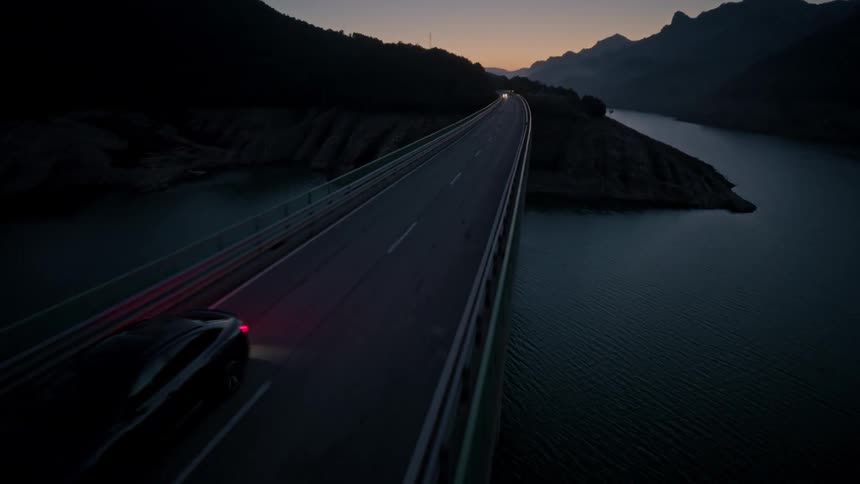 A lone car with its headlights on drives across a long bridge spanning a dark body of water surrounded by mountains at dusk