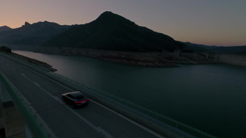 Black car drives on bridge over lake at dusk with mountains behind