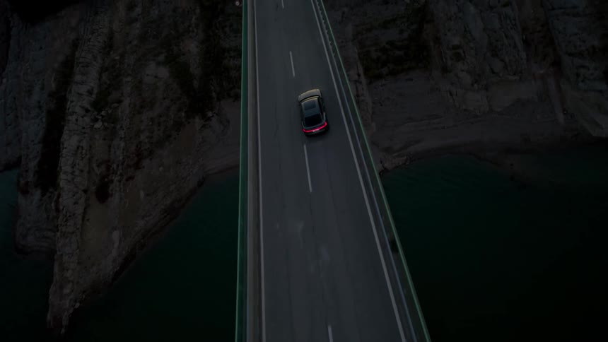 An aerial shot captures a black car driving across a long bridge spanning a dark body of water flanked by mountains at morning