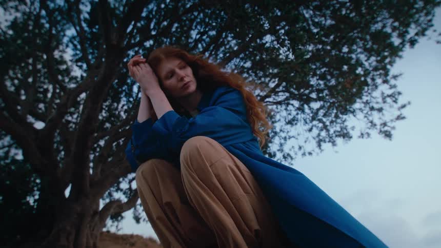 A woman with red hair wearing a blue jacket squats beside a tree on a cloudy day looking sad as a soft wind blows her clothes and hair
