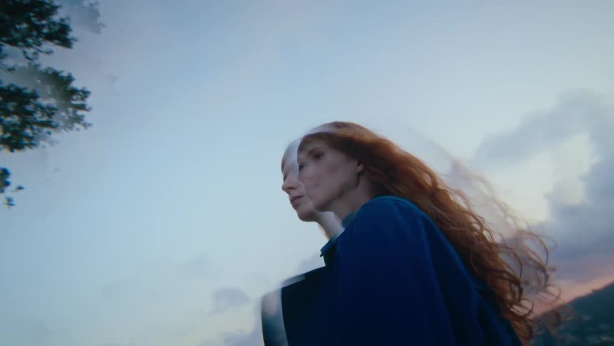 A red haired woman walking with her hair blowing in the wind against a vast cloudy sky at dusk