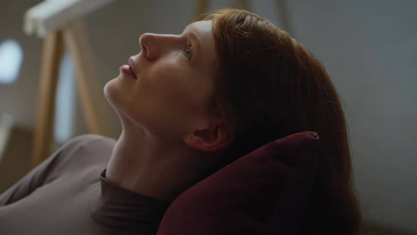 A red haired woman lies with her head on a pillow opening her eyes and looking upwards with a contemplative expression