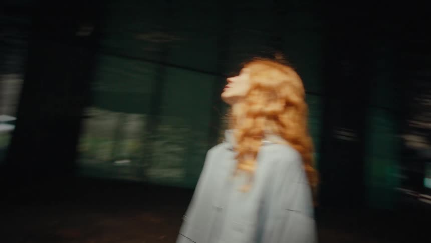 A woman with long red hair stands in a room as the camera spins around her chaotically